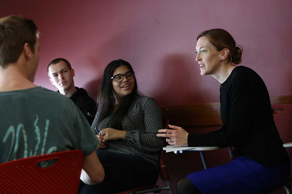 Female faculty member, sitting at table with students, speaking to them.