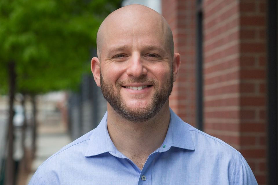 Head shot of a smiling man with a beard wearing a light-blue shirt, taken outside near some trees and a brick building. 